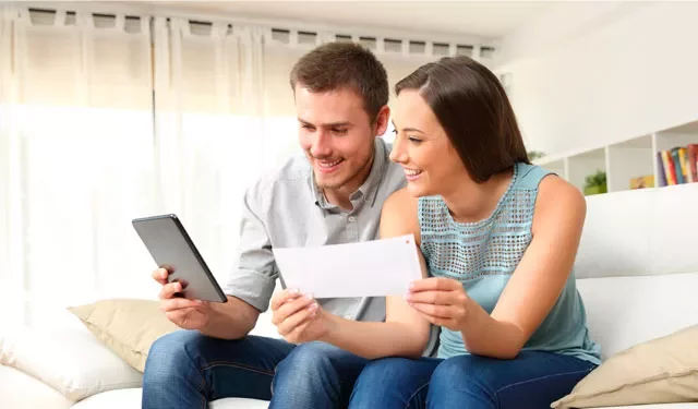 Two people on a couch looking at a tablet and a piece of paper, smiling.