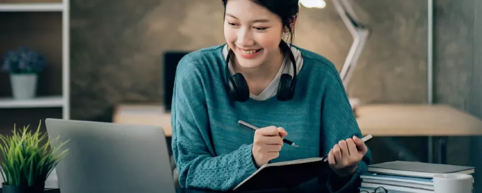 Person smiling at a laptop while taking notes at a desk.