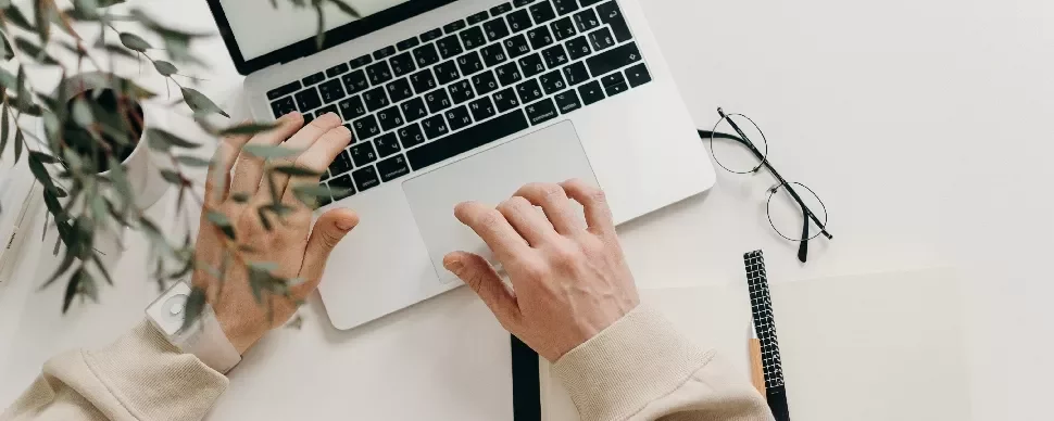Person using a laptop with a notebook and glasses nearby on a desk.