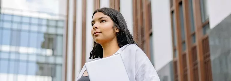 Person holding a laptop, standing in front of modern building.