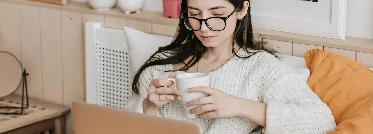 Person in a cozy room, reading on a laptop and holding a cup.