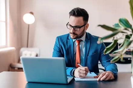Person in blue suit works at laptop, jotting notes in a notebook.