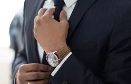 Person adjusting a tie while wearing a navy suit and a watch with a brown leather strap.